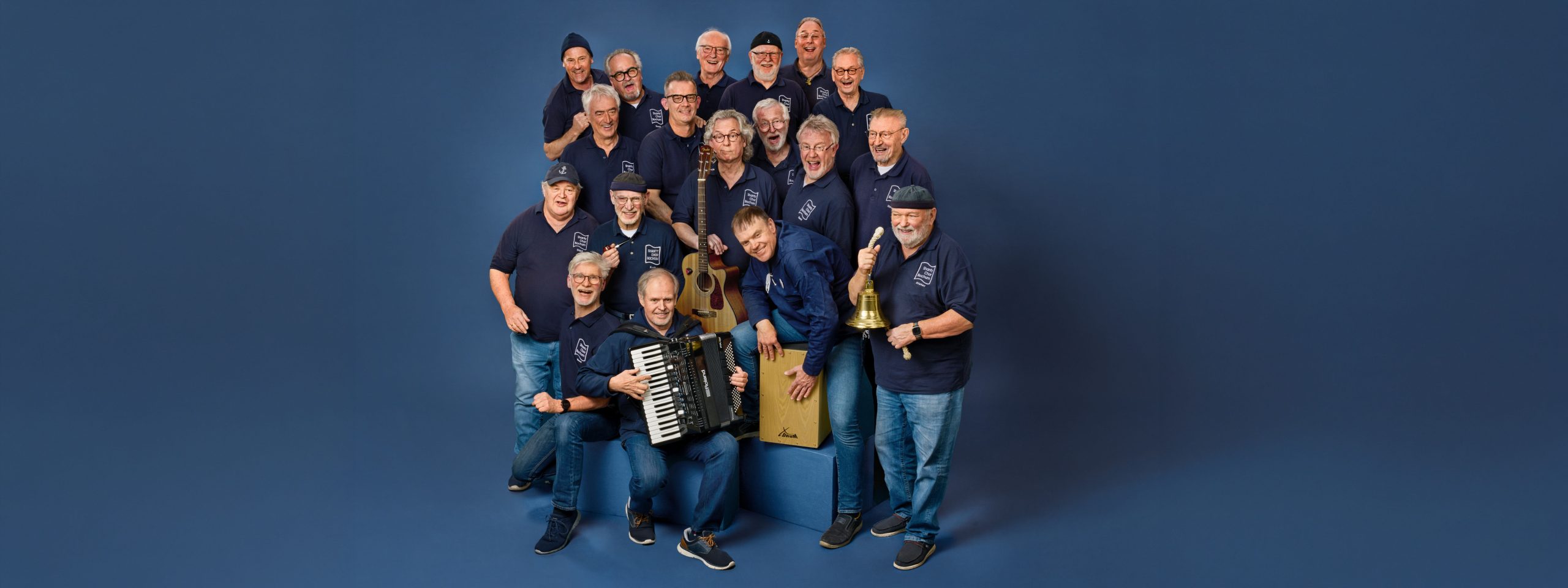 Eine Gruppe von 18 Männern steht zusammen. Einige halten ein Akkordeon, eine Gitarre, das Instrument Cajón, eine Schiffsglocke und eine Tabakpfeife in den Händen. Sie lächeln.