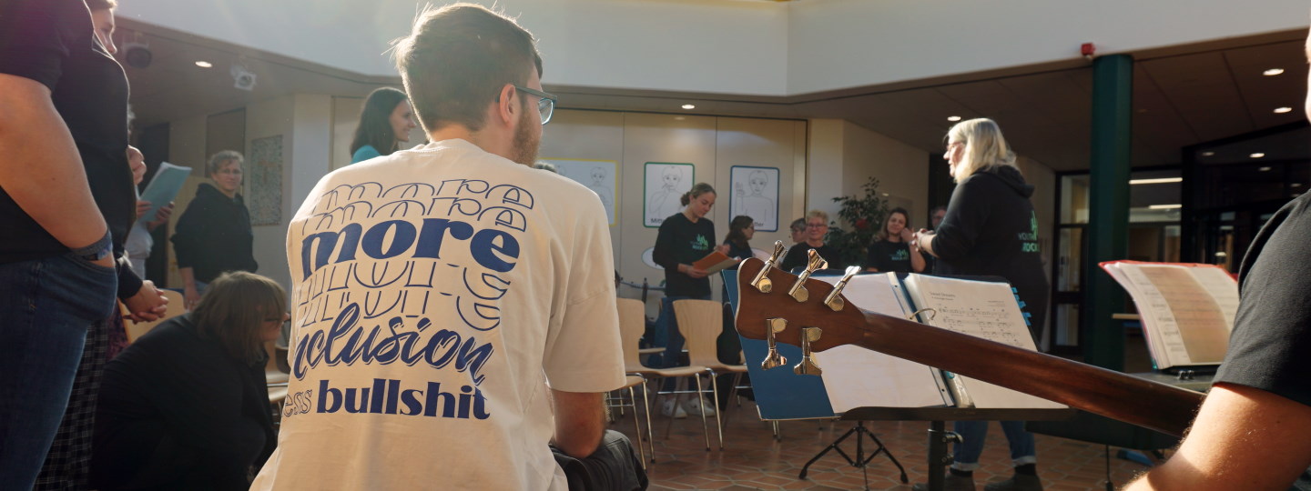 In einer Schulaula stehen Menschen im Kreis um eine Chorleiterin, die Atmosphäre ist entspannt. Ein junger Mann im Vordergrund trägt ein T-Shirt, auf dem mehr Inklusion gefordert wird.