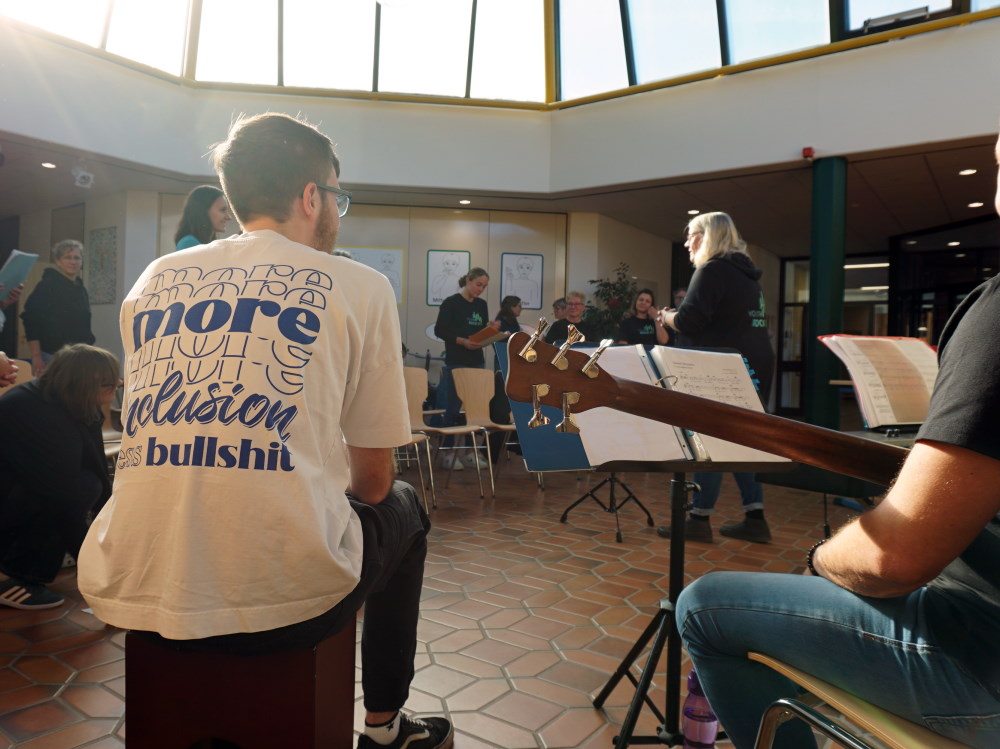 In einer Schulaula stehen Menschen im Kreis um eine Chorleiterin, die Atmosphäre ist entspannt. Ein junger Mann im Vordergrund trägt ein T-Shirt, auf dem mehr Inklusion gefordert wird.