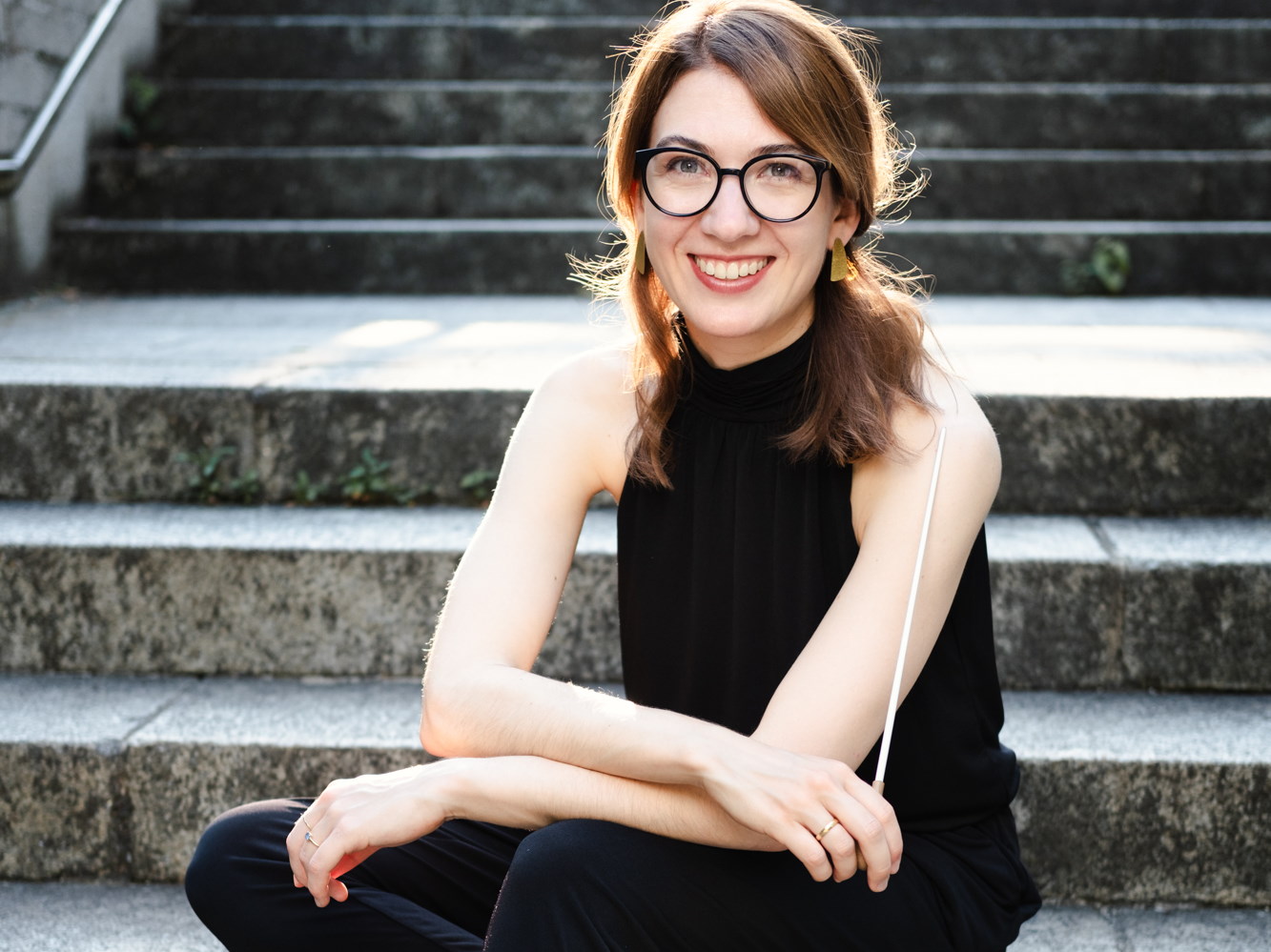 Die Dirigentin Katharina Morin, mit schwarzem Brillengestellt und goldenen Ohrringen, schaut lächelnd in die Kamera. Im Hintergrund sind undeutlich Stufen zu erkennen.