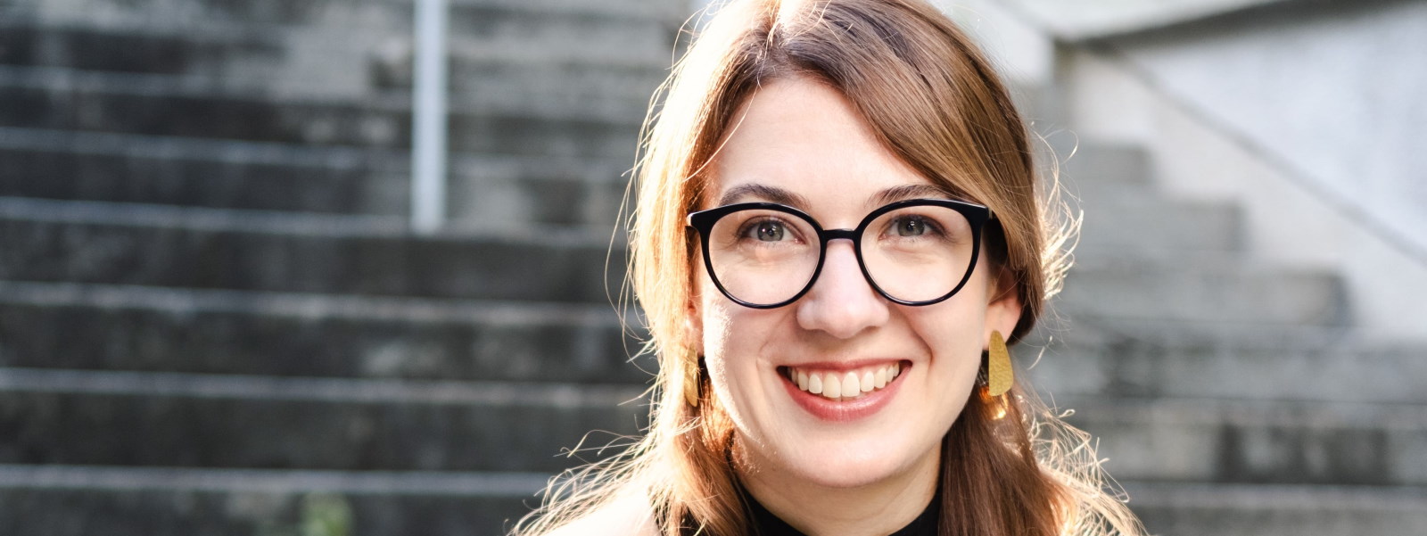 Die Dirigentin Katharina Morin, mit schwarzem Brillengestellt und goldenen Ohrringen, schaut lächelnd in die Kamera. Im Hintergrund sind undeutlich Stufen zu erkennen.