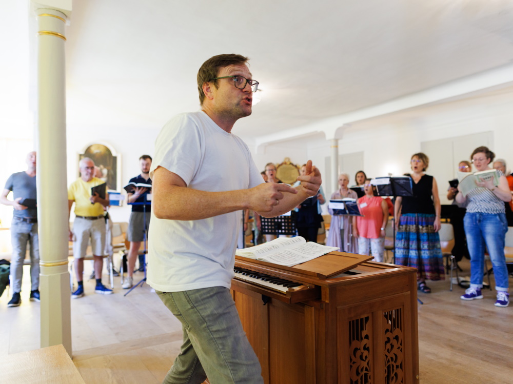 Benno Schachtner, in Jeans und weißem T-Shirt an einer Truhenorgel stehend, dirigiert einen gemischten Chor, der um ihn herum angeordnet steht.