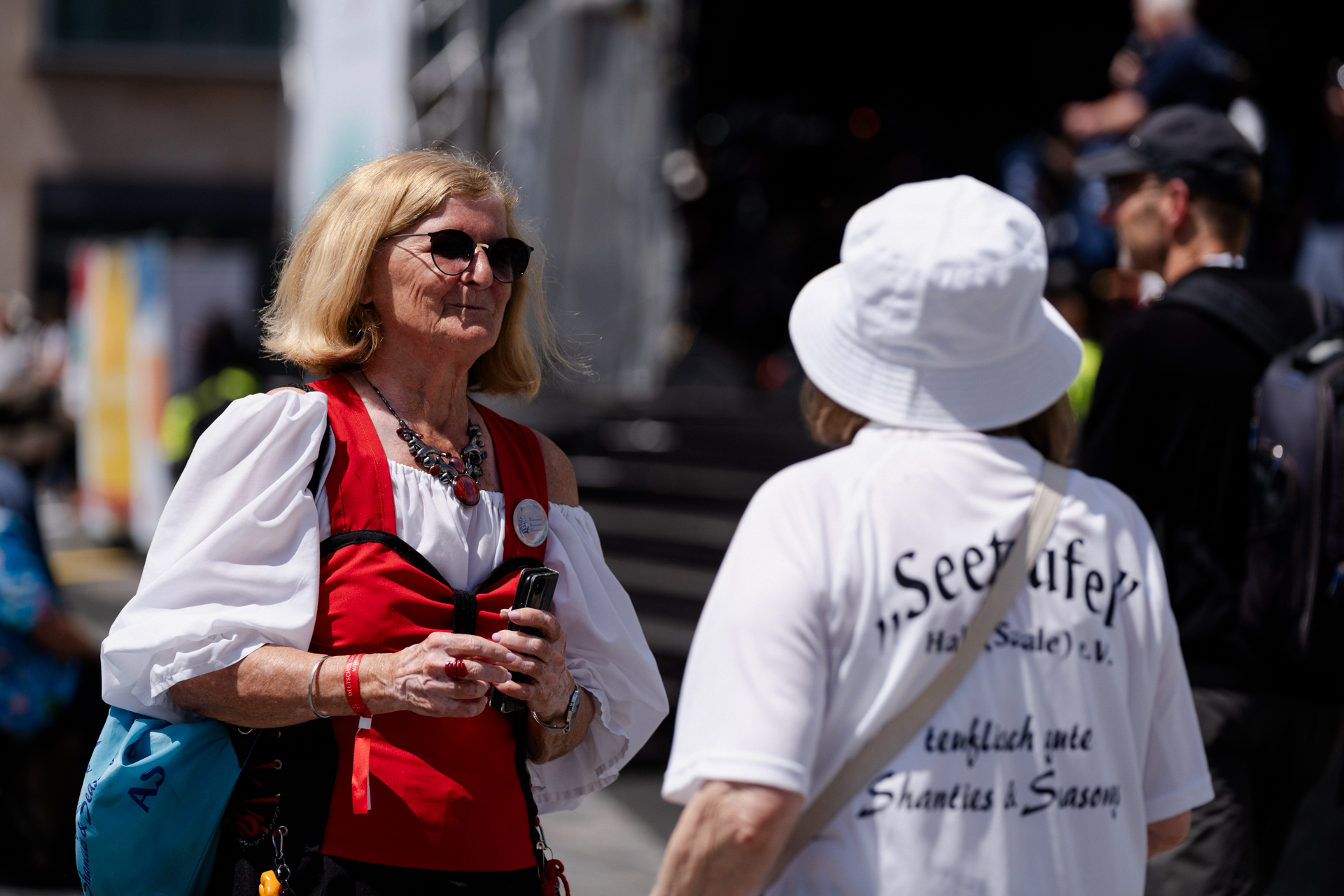 Eine Frau von vorne im roten Kleid der Seeteufel. Eine Person von hinten mit einem T-Shirt auf dem Seeteugel, teuflisch gute Shanties und Seasongs steht.