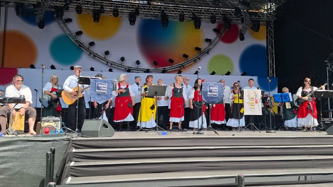 Etwa 20 Männer und Frauen stehen auf einer Bühne. Die Frauen tragen rote und gelbe Röcke, die Männer weiße Hemden und dunkle Hosen. Einige Menschen spielen Gitarre und trommelt auf einem Cajon.