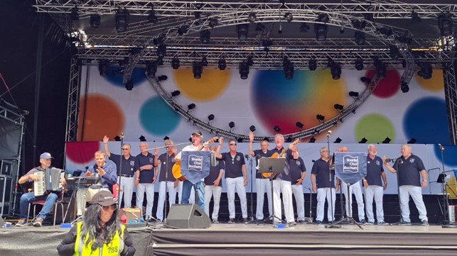 Etwa 15 Männer in weißen Hosen und dunkelblauen T-Shirts singen auf einer Bühne. Ein Mann spielt Akkordeon, ein Mann Klavier, zwei Männer Gitarre. Im Hintergrund sind Lichter und ein Vorhang mit bunten Kugeln.