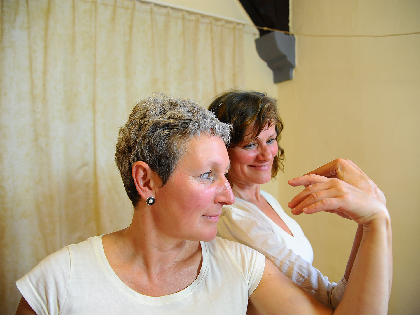 Zwei Frauen, die Schulter an Schulter stehen, haben zur selben Seite jeweils einen Arm gehoben und beobachten die oberhalb des Unterarms locker ruhende Hand. Beide lächeln.