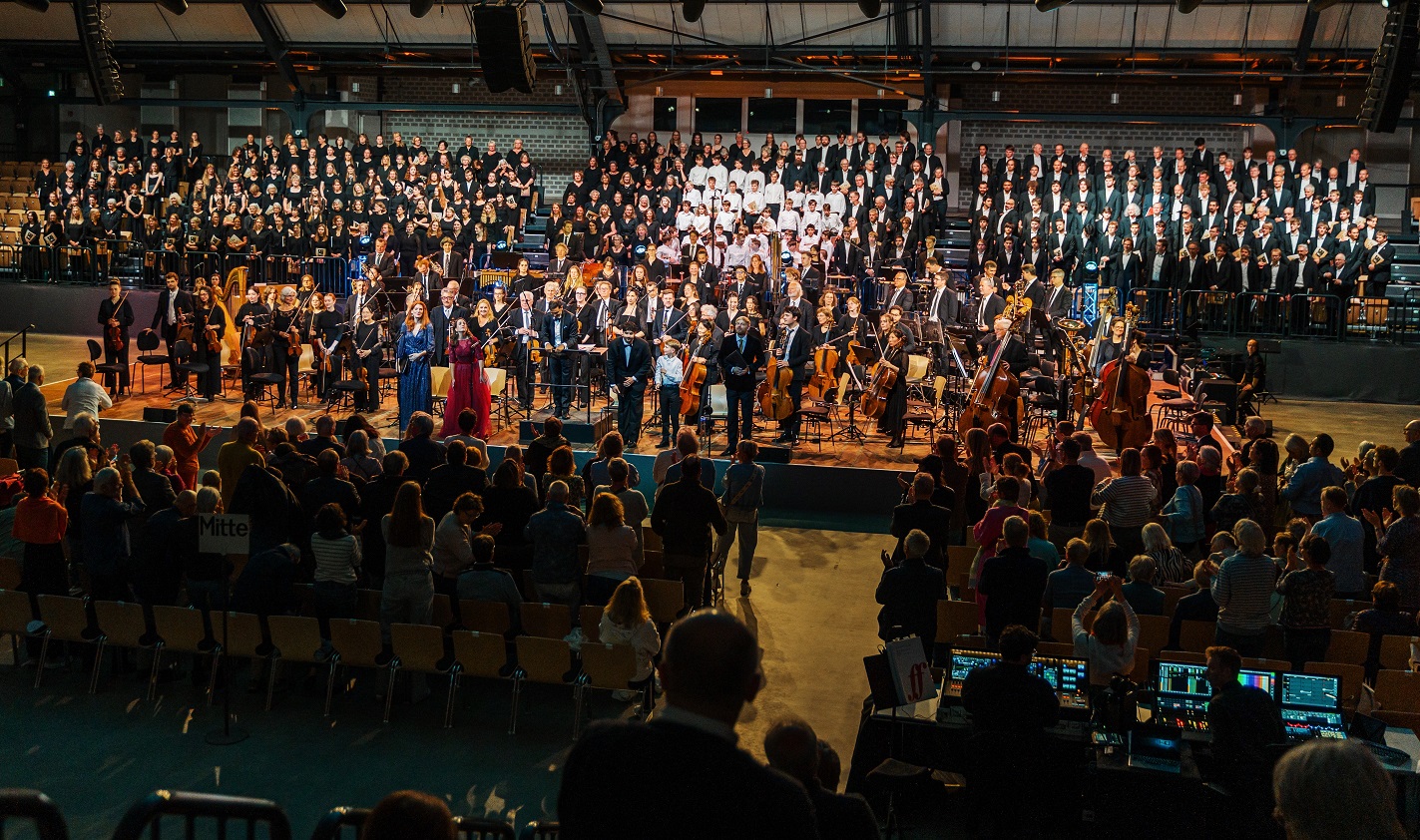Hunderte Sängerinnen und Sänger sowie Menschen mit Musikinstrumenten stehen auf einer Bühne