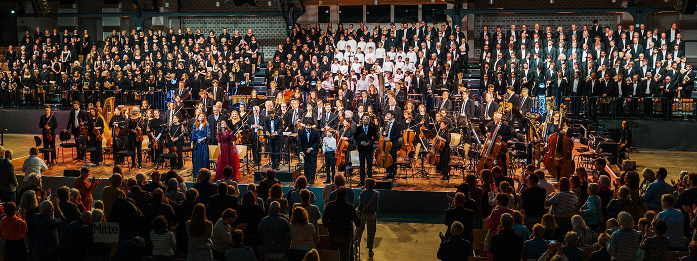 Hunderte Sängerinnen und Sänger sowie Menschen mit Musikinstrumenten stehen auf einer Bühne. Der Publikumsraum ist gefüllt mit Zuschauenden.
